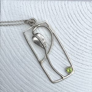 The handmade Sterling Silver necklace features a rectangular pendant showcasing a minimalist design of a leaf and stem with flowing lines. A small, round, green peridot is set into the bottom right corner of the frame. The pendant is attached to a delicate silver chain.